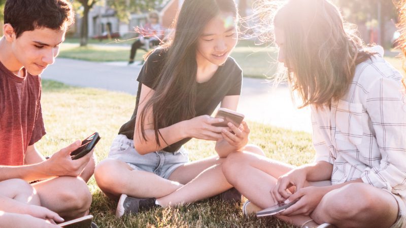 Group of young teenagers sitting outside in a circle showing each other images on their phones