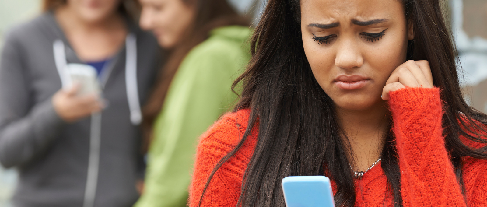 Teen looking sadly at phone while two other teens laugh in the background, indicating that she is experiencing cyberbullying.