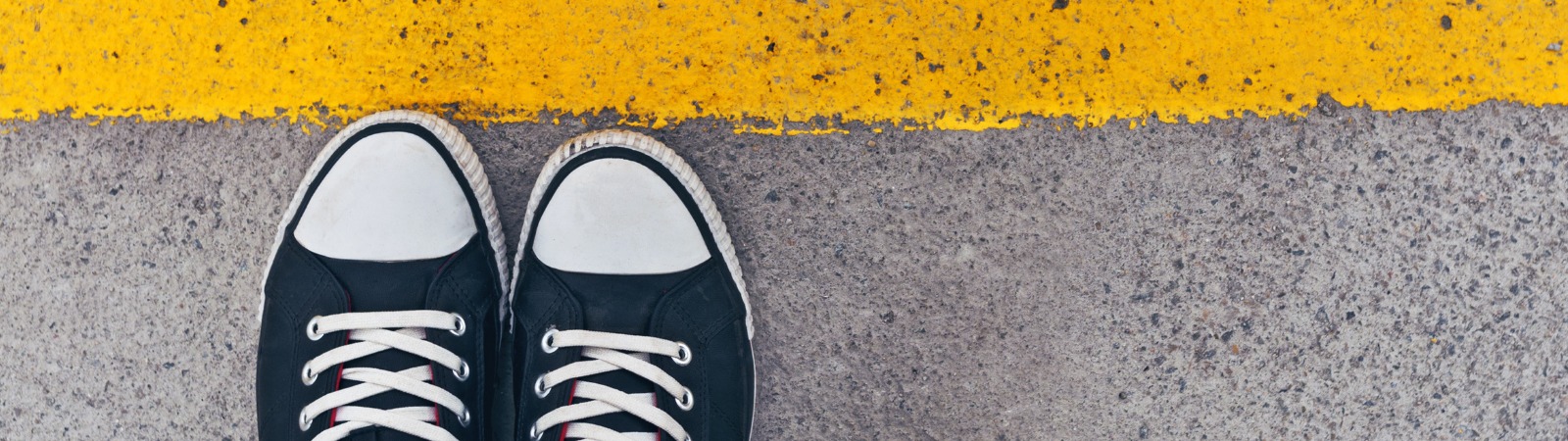 Pair of shoes stepping right up to a boundary illustrated by a yellow line painted on a sidewalk.