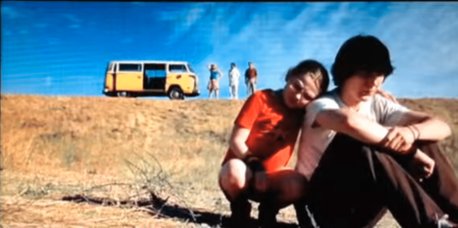 Scene from the movie Little Miss Sunshine where a young girl dressed in red is comforting her teenage brother. They are sitting on the ground at the bottom of the hill with the family van and parents in the background.