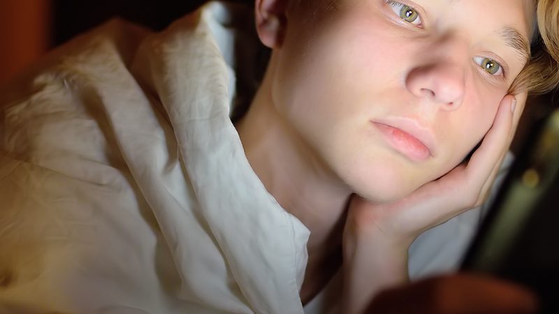 White teenager laying on bed looking at their cell phone. Their face is illuminated by the glow of the phone.