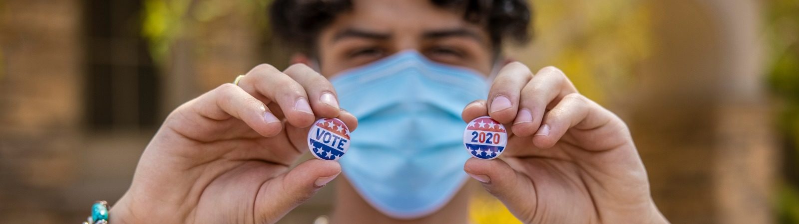 Masked teenager holding up two pins that say VOTE and 2020