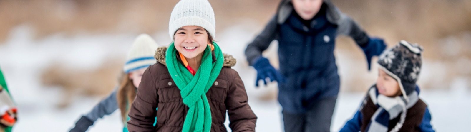 Two children in winter clothes running across a field with a parent following behind them.